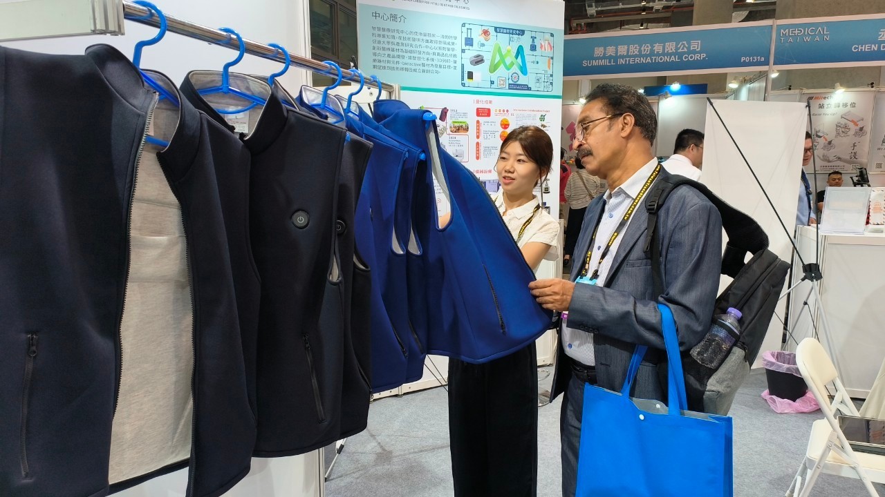這張照片拍攝於展覽會場，畫面中一位女性正在向男性參觀者介紹掛在架上的智慧照護背心。這些背心有黑色與藍色款式，並帶有按鈕與拉鍊，可能具有智慧科技功能，例如體溫調節或健康監測。  背景展示板上有關於展出的簡介與圖示，標示出這項產品的功能與應用場景。男性參觀者手持藍色購物袋，並背著背包，顯示他可能正在參觀不同攤位，尋找適合的技術或產品。整體氛圍專業，呈現出展覽交流與產品推廣的情境。
