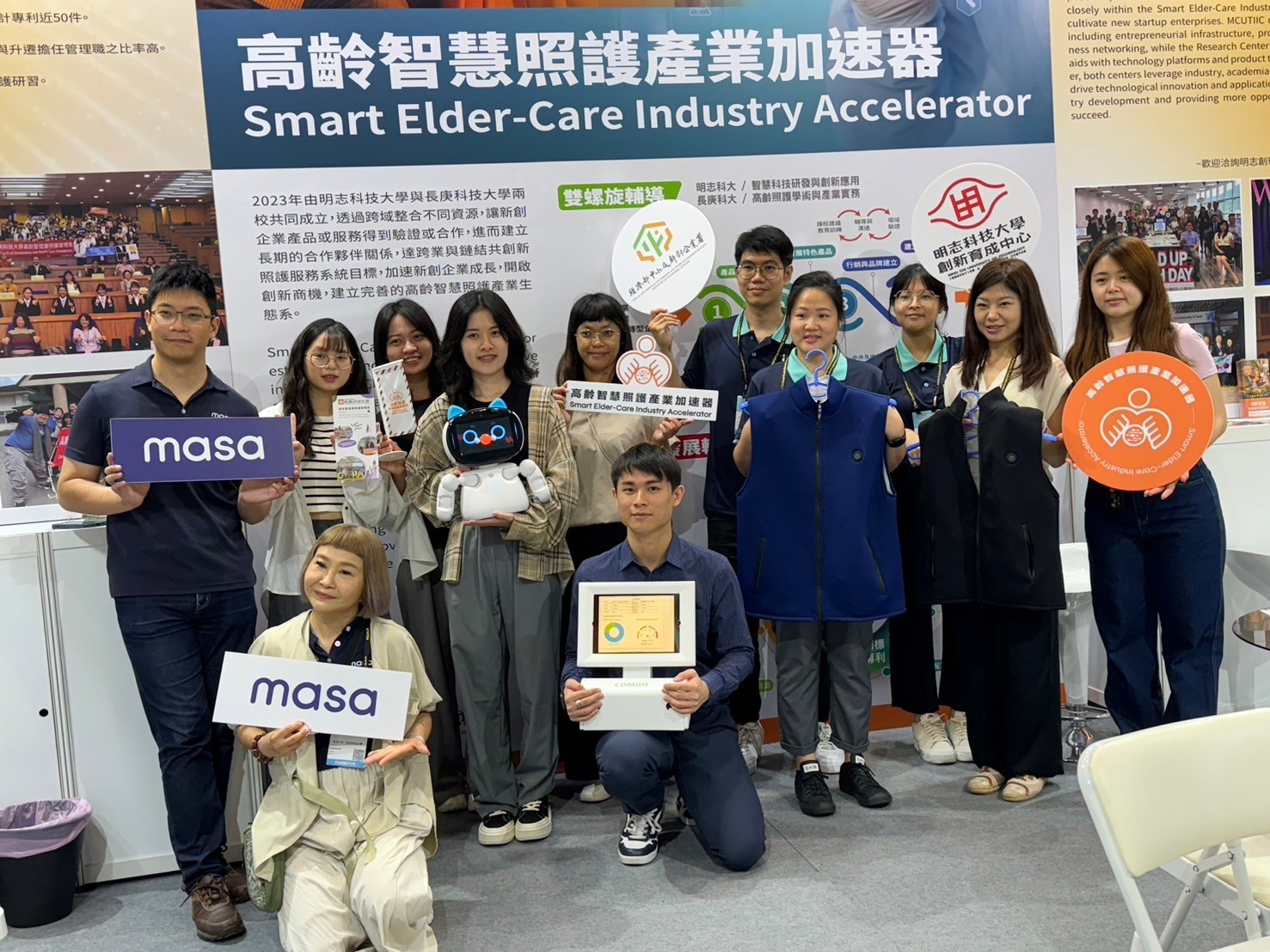 這張照片拍攝於一個展覽或活動現場，背景看板標示「高齡智慧照護產業加速器」（Smart Elder-Care Industry Accelerator），顯示活動與高齡照護和智慧科技相關。  照片中的人員約有十多位，皆面帶笑容，展示相關產品，包括智慧型機器人、智慧照護背心、宣傳手冊和標誌牌等。其中，有人手持「masa」標誌，可能是參展企業或品牌名稱。現場氛圍活躍，參與者來自不同機構，可能包括學界、產業界或創新團隊，共同推動高齡照護產業的發展。