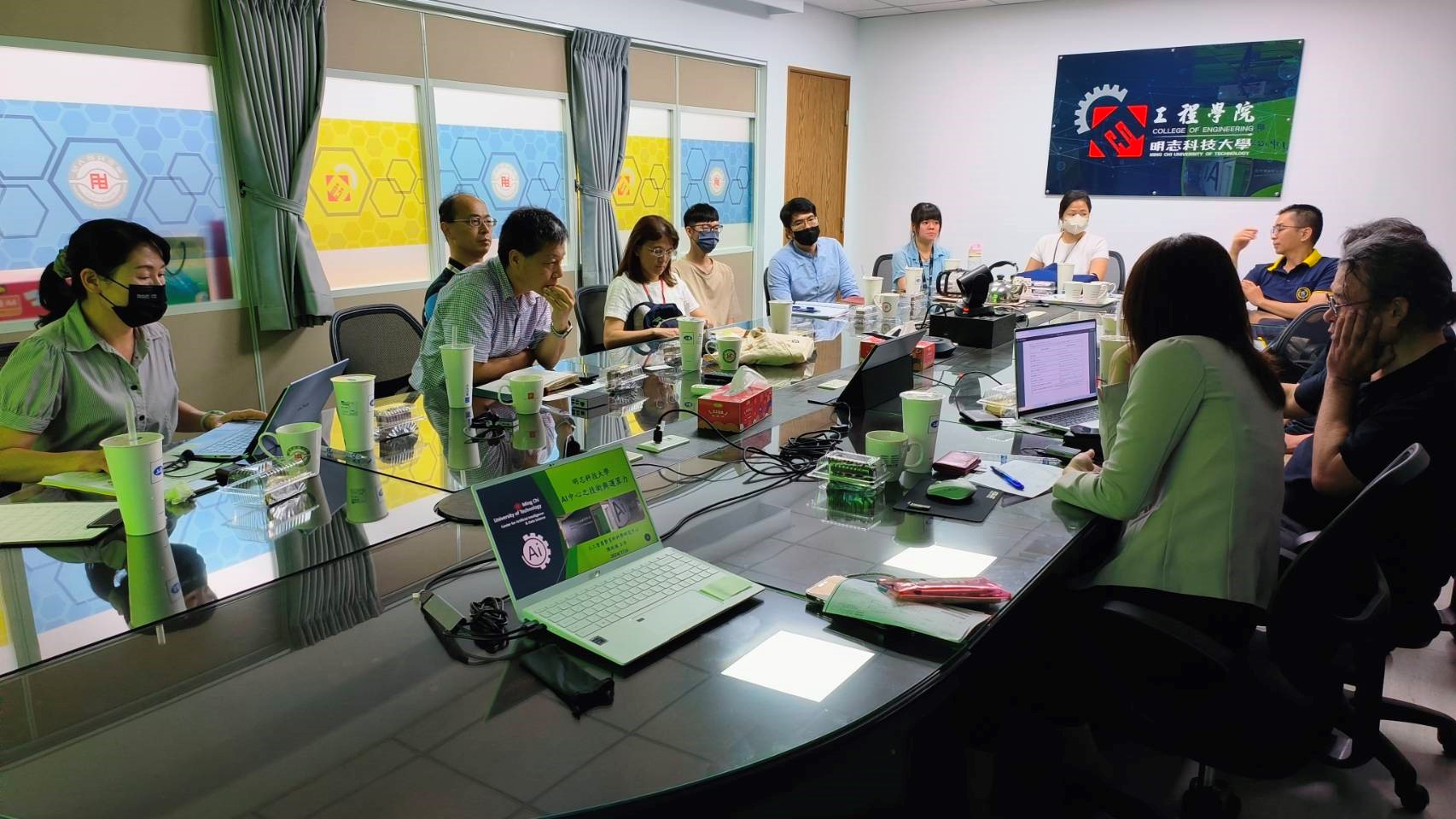 這張照片顯示了一場會議，與會者約有十多位，坐在長桌旁進行討論。現場環境為會議室，牆上有「眾志科技大學 工程學院」的標誌，顯示會議可能與技術或工程相關。桌上擺滿了筆記型電腦、文件、飲料和其他辦公用品，顯示出專業的會議氛圍。部分與會者佩戴口罩，可能是為了防疫考量，並且大家專注於討論或記錄資訊。前方的筆電畫面顯示「AI+工業生產力」，推測會議內容可能與人工智慧及工業應用相關。