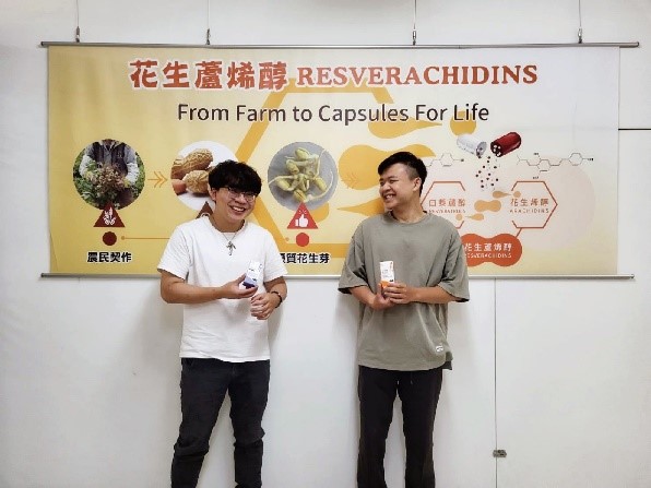 這張圖片中可以看到兩位年輕男性站在一面宣傳布條前，彼此微笑交談，氣氛輕鬆愉快。布條上寫著「花生蕊烯醇 RESVERACHIDINS」以及「From Farm to Capsules For Life」，並搭配相關圖示，說明產品從農民契作到萃取製成膠囊的過程。這張照片應該是與某項健康食品或農業加工產品的推廣活動有關。